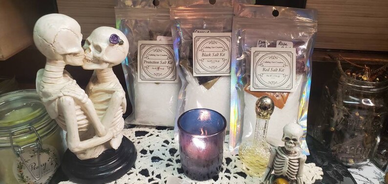 May include: Three clear plastic bags containing salt kits for various purposes. The bags are labeled "Protection Salt Kit", "Black Salt Kit", and "Red Salt Kit". The kits are displayed on a white lace tablecloth with a small glass jar of green liquid, a small glass bottle with a gold top, and a small glass candle holder with a purple candle.