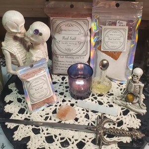 May include: A white crocheted doily sits on a black surface with a silver sword, a small pink Himalayan salt rock, a purple candle holder, a clear glass bottle with a silver top, and two small bags of red salt. The bags are labeled "Red Salt" and "Red Salt Kit".  Two white skeleton figurines are on either side of the doily.