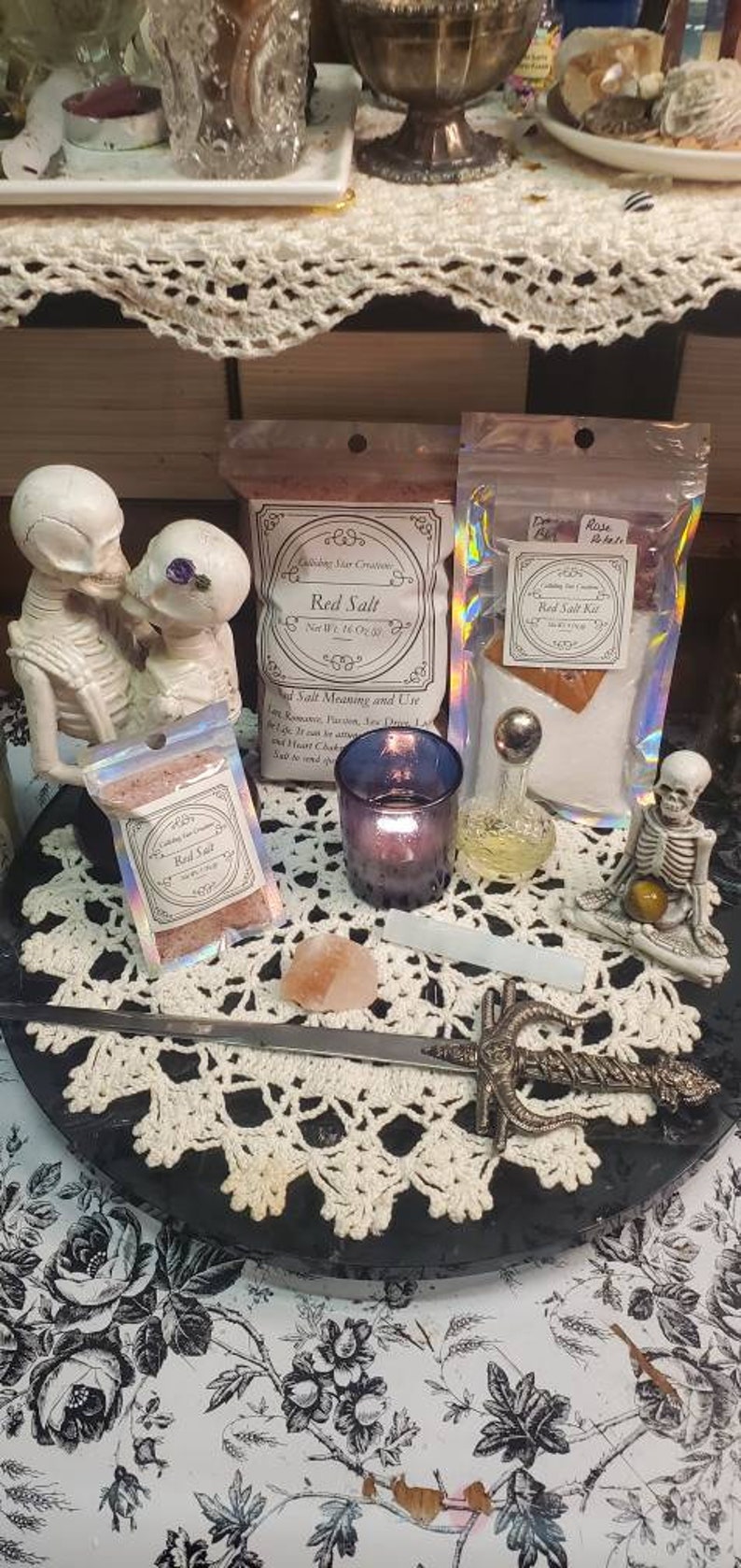 May include: A white crocheted doily sits on a black surface with a silver sword, a small pink Himalayan salt rock, a purple candle holder, a clear glass bottle with a silver top, and two small bags of red salt. The bags are labeled "Red Salt" and "Red Salt Kit".  Two white skeleton figurines are on either side of the doily.