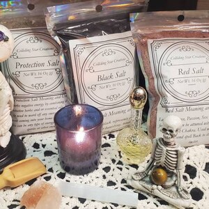 May include: Three bags of salt labeled "Protection Salt", "Black Salt", and "Red Salt" with a description of the salt's meaning and use.  The bags are on a white lace tablecloth with a skeleton figurine, a candle, and a glass bottle.
