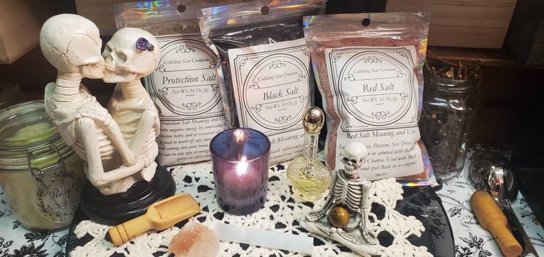May include: Three bags of salt labeled "Protection Salt", "Black Salt", and "Red Salt" with a description of the salt's meaning and use.  The bags are on a white lace tablecloth with a skeleton figurine, a candle, and a glass bottle.