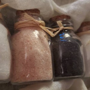 May include: Five small glass bottles with cork tops and twine tied around the necks. The bottles are filled with different colored salts, including white, pink, black, and gray.