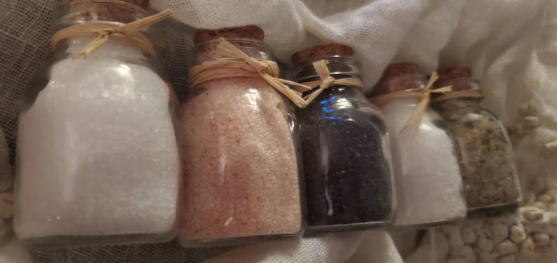 May include: Five small glass bottles with cork tops and twine tied around the necks. The bottles are filled with different colored salts, including white, pink, black, and gray.