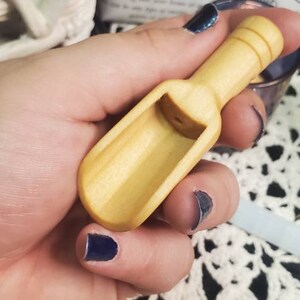 May include: A small wooden scoop with a rounded handle. The scoop is light brown and has a smooth finish. The scoop is being held in a hand with dark blue nail polish.