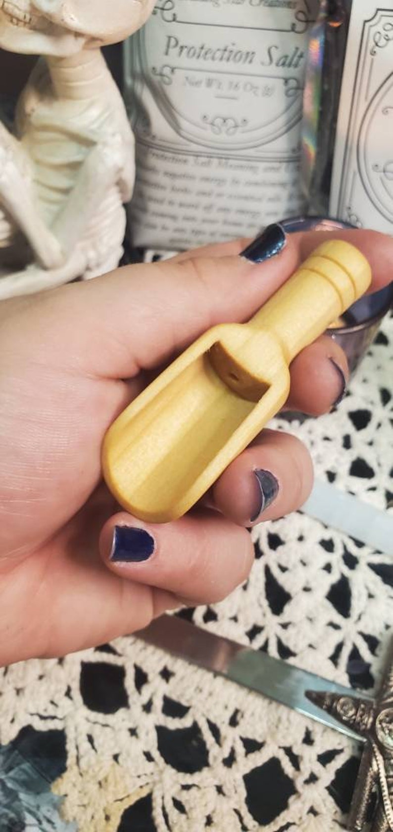 May include: A small wooden scoop with a rounded handle. The scoop is light brown and has a smooth finish. The scoop is being held in a hand with dark blue nail polish.