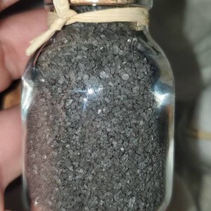 May include: A small glass jar with a cork lid filled with black salt crystals. The jar is tied with a natural fiber ribbon.