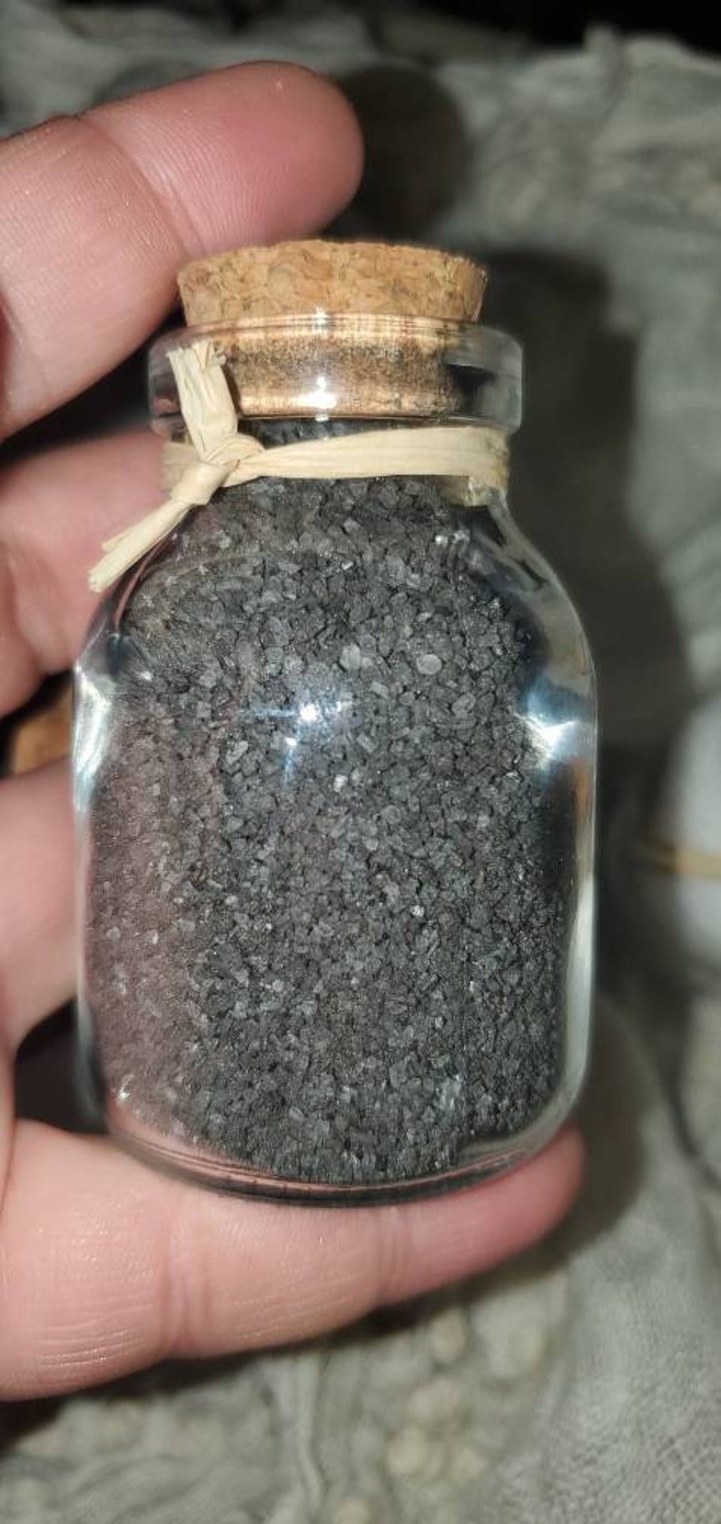 May include: A small glass jar with a cork lid filled with black salt crystals. The jar is tied with a natural fiber ribbon.