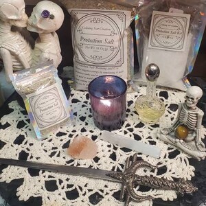 May include: A white crocheted doily sits on a black surface with a silver dagger, a small pink crystal, a clear glass bottle with a silver stopper, a purple candle, and two small bags of protection salt. The bags are labeled "Protection Salt" and "Protection Salt Kit". Two white skeleton figurines are in the background.