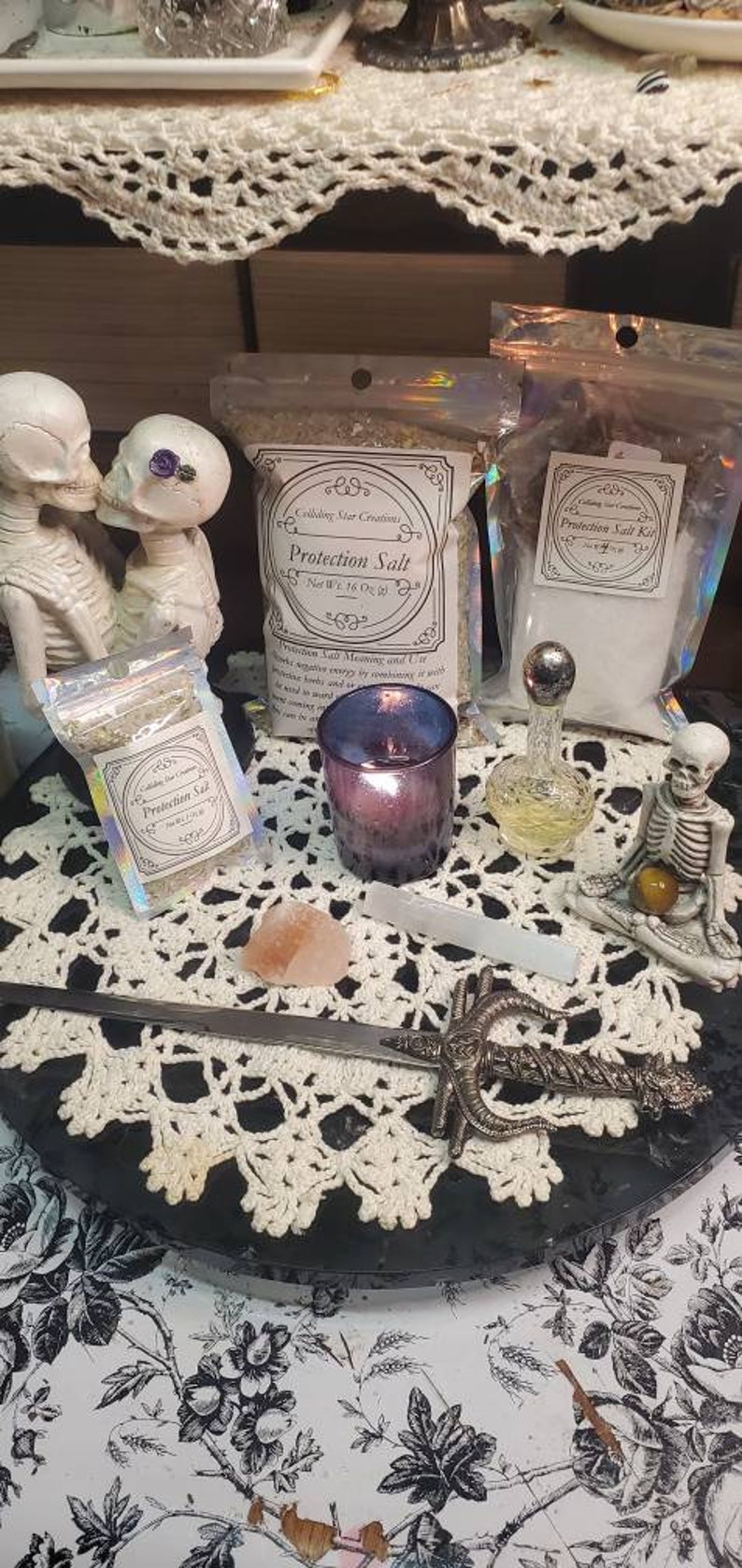 May include: A white crocheted doily sits on a black surface with a silver dagger, a small pink crystal, a clear glass bottle with a silver stopper, a purple candle, and two small bags of protection salt. The bags are labeled "Protection Salt" and "Protection Salt Kit". Two white skeleton figurines are in the background.