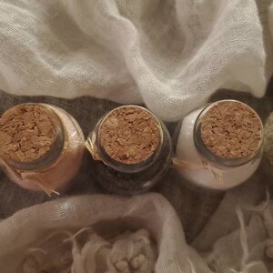 May include: Five small glass jars with cork lids, filled with different colored powders. The jars are arranged in a row on a white fabric background.