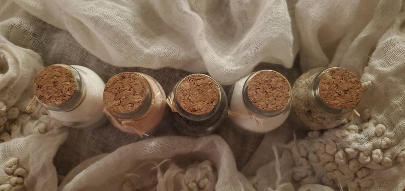 May include: Five small glass jars with cork lids, filled with different colored powders. The jars are arranged in a row on a white fabric background.