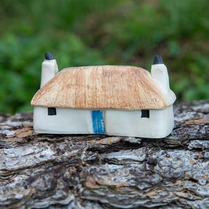 May include: A small white ceramic cottage with a thatched roof and a blue door. The cottage is sitting on a piece of tree bark.