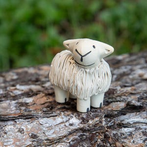 May include: A white ceramic sheep figurine with a fluffy texture and black eyes. The sheep is standing on a brown tree bark surface.