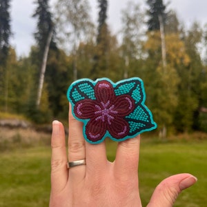 May include: A turquoise and maroon beaded flower ring. The ring is on a person's finger.
