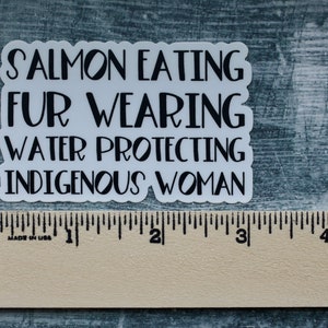Peut inclure: Autocollant blanc avec du texte noir qui dit "Salmon Eating Fur Wearing Water Protecting Indigenous Woman". L'autocollant est plac&eacute; sur une r&egrave;gle en bois.