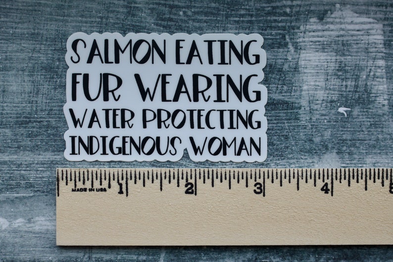 Peut inclure: Autocollant blanc avec du texte noir qui dit "Salmon Eating Fur Wearing Water Protecting Indigenous Woman". L'autocollant est plac&eacute; sur une r&egrave;gle en bois.