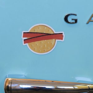 May include: A blue refrigerator with a silver handle and a magnet of a cracker with two slices of meat on top. The magnet says "G" on the top right corner.