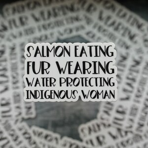Peut inclure: Un autocollant blanc avec du texte noir qui dit "Salmon Eating Fur Wearing Water Protecting Indigenous Woman". L'autocollant est sur un fond de nombreux autres autocollants avec un texte similaire.