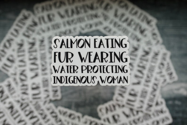 Peut inclure: Un autocollant blanc avec du texte noir qui dit "Salmon Eating Fur Wearing Water Protecting Indigenous Woman". L'autocollant est sur un fond de nombreux autres autocollants avec un texte similaire.