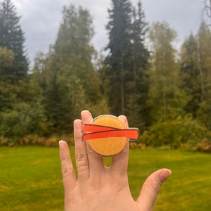 May include: A yellow and orange ring with two red stripes, resembling a hamburger, worn on a finger.