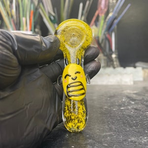 May include: A yellow glass pipe with black speckles and a cartoon face with a surprised expression. The pipe is shaped like a spoon with a rounded bowl.