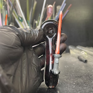 May include: A dark glass pipe with a red and white lightsaber design. The pipe is held in a gloved hand.