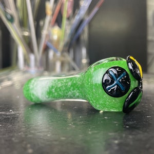 May include: Green glass pipe with black and yellow accents. The pipe has a speckled green finish and features three black buttons with white crosses.