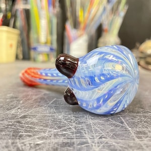 May include: A blue and white glass pipe with red accents and dark brown details. The pipe has a swirling pattern and is made of clear glass with blue and white swirls.