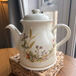 May include: Cream-colored ceramic teapot with a lid, spout, and handle. The teapot is decorated with a floral and berry design in shades of green, brown, and purple. It sits on a woven wicker coaster on a wooden table.