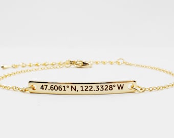 May include: A gold chain bracelet with a thin, rectangular tag engraved with the coordinates "47.6061° N, 122.3328° W".