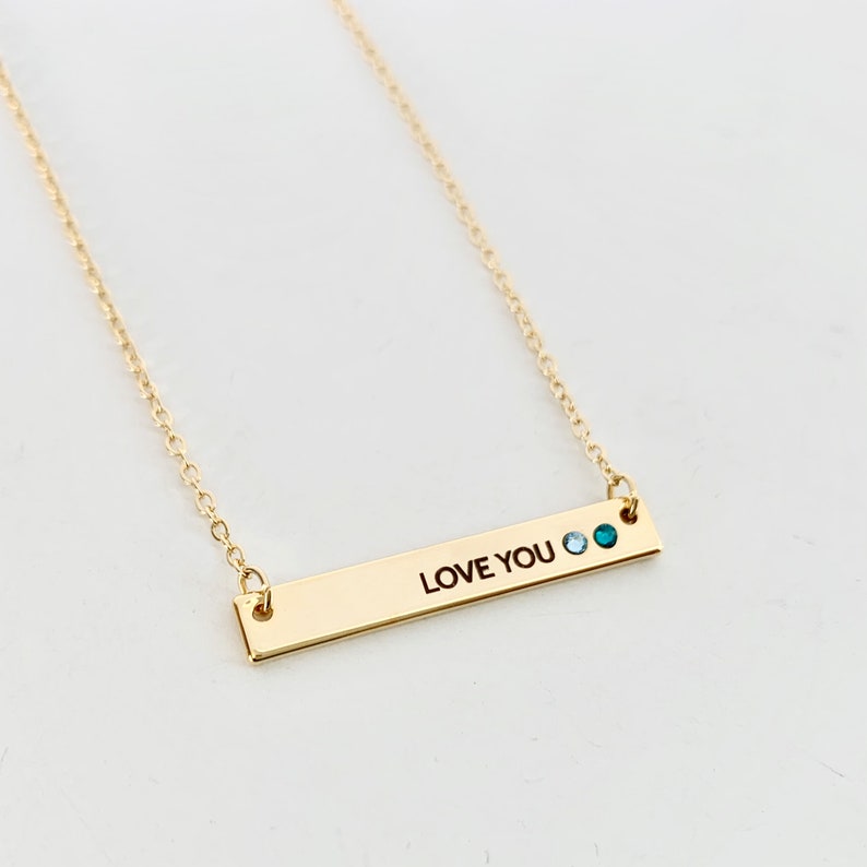 May include: Gold bar necklace with the words "LOVE YOU" engraved on it. The necklace has two small blue gemstones.