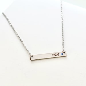 May include: A silver bar necklace with the name "LUCAS" engraved on it. A small blue gemstone is set on the bar.