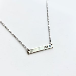 May include: Silver necklace with a bar pendant engraved with the name "LUCAS" and a small blue gemstone.