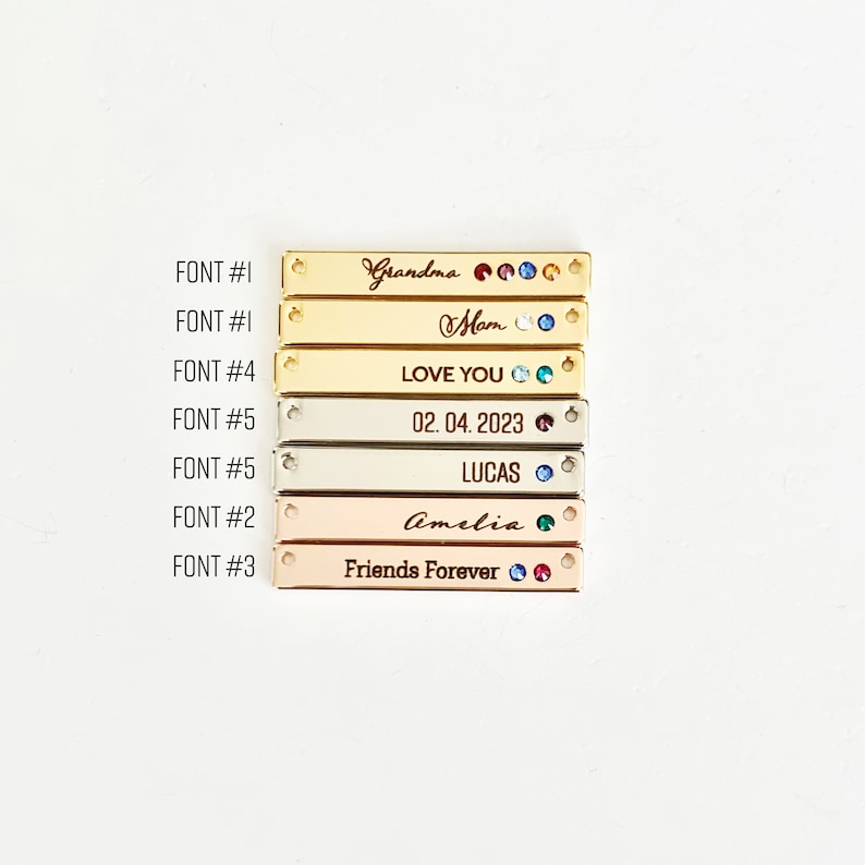 May include: A set of seven gold, silver, and rose gold bar pendants with various engraved text and rhinestones. The text includes "Grandma", "Mom", "Love You", "02.04.2023", "Lucas", "Amelia", and "Friends Forever".