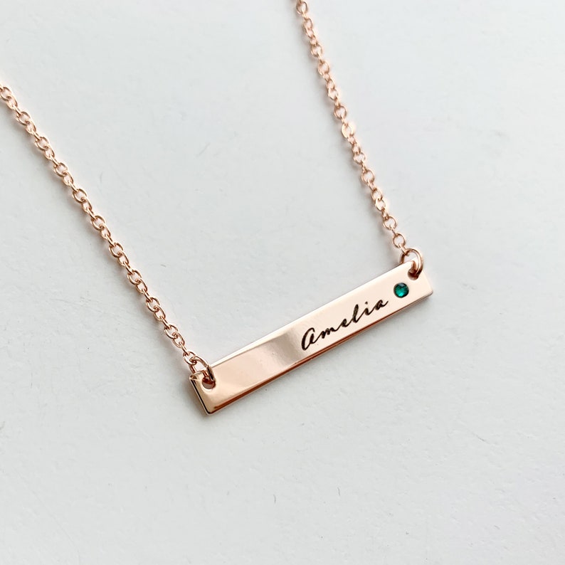 May include: A rose gold bar necklace with the name "Amelia" engraved on it. A small green gemstone is set on the bar.