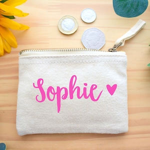 May include: A small, white canvas pouch with a zipper closure. The pouch has the name "Sophie" written in pink cursive with a pink heart next to it.