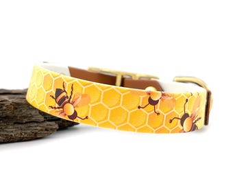 Puede incluir: Un collar amarillo para perros con un patrón de panal y abejas de dibujos animados. El collar tiene un respaldo de cuero marrón y herrajes dorados. El collar está sobre un trozo de madera.