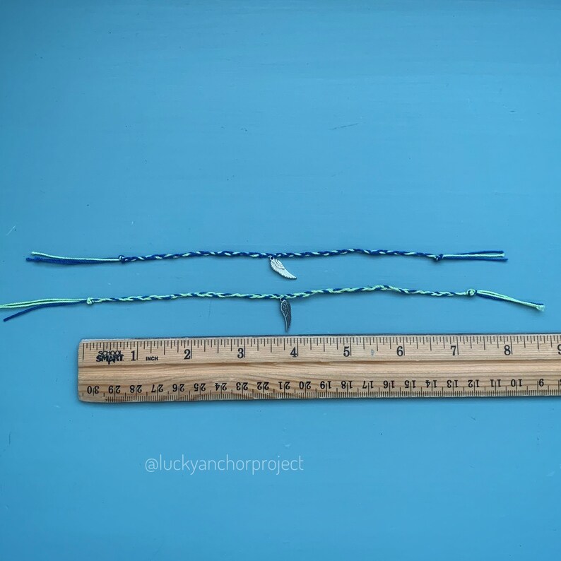 May include: Two friendship bracelets with a silver feather charm. The bracelets are made with blue and green string. The bracelets are laid out on a blue surface with a ruler underneath.