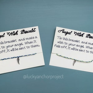 May include: Two white cards with black text and a blue and green string bracelet with a silver feather charm. The text on the cards reads "Angel Wish Bracelet" and "Tie this bracelet and make a wish to your angel. When it falls off, it will be sent to them." The text "@luckyanchorproject" is at the bottom of the cards.