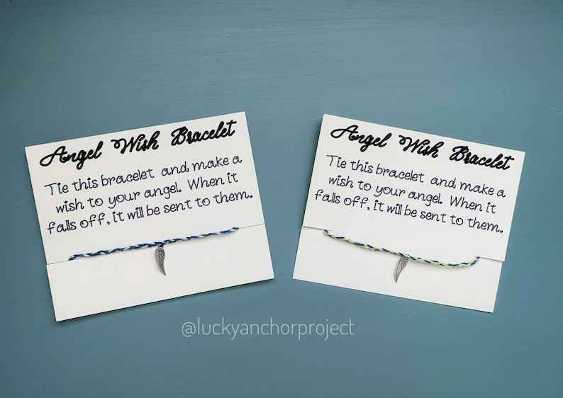 May include: Two white cards with black text and a blue and green string bracelet with a silver feather charm. The text on the cards reads "Angel Wish Bracelet" and "Tie this bracelet and make a wish to your angel. When it falls off, it will be sent to them." The text "@luckyanchorproject" is at the bottom of the cards.