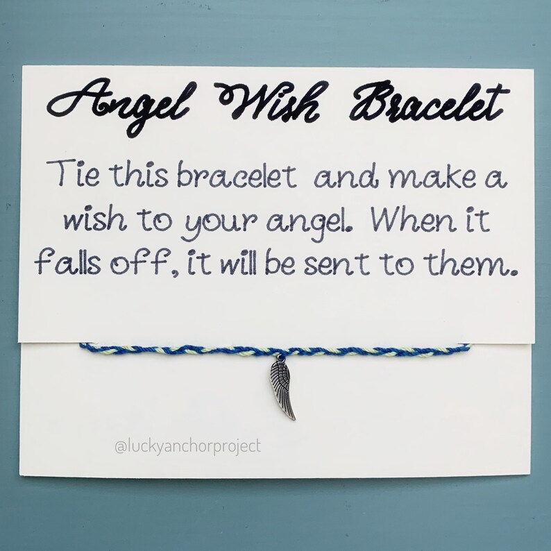 May include: A white card with blue and green string tied around it. The string has a silver angel wing charm attached. The card reads "Angel Wish Bracelet" and "Tie this bracelet and make a wish to your angel. When it falls off, it will be sent to them." The card also has the text "@luckyanchorproject" at the bottom.
