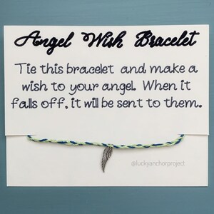 May include: A white card with black text that reads "Angel Wish Bracelet". The text continues, "Tie this bracelet and make a wish to your angel. When it falls off, it will be sent to them." A blue and green string bracelet with a silver angel wing charm is attached to the card. The text "@luckyanchorproject" is at the bottom of the card.