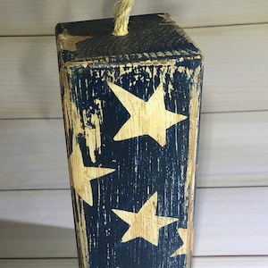 May include: A distressed wooden block painted navy blue with cream-colored stars. A natural rope is attached to the top. The block has a rustic, vintage appearance, with visible wood grain and a worn finish. The design evokes a patriotic theme.