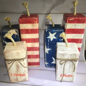 May include: A collection of rustic, wooden firecracker decorations in red, white, and blue. Some feature the American flag design, while others have a distressed, vintage look. Each has a jute rope fuse and is tied with twine.