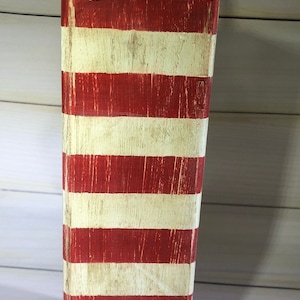 May include: A rectangular wooden decorative buoy painted with alternating red and off-white horizontal stripes. A natural rope is attached to the top. The buoy has a distressed, vintage appearance, suggesting nautical decor.