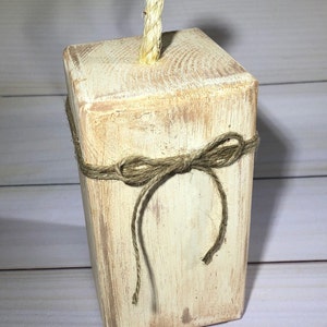 May include: A rustic, rectangular wooden block with a distressed, off-white finish. A natural rope bow is tied around the middle, and a twisted rope extends from the top. The block has visible wood grain.