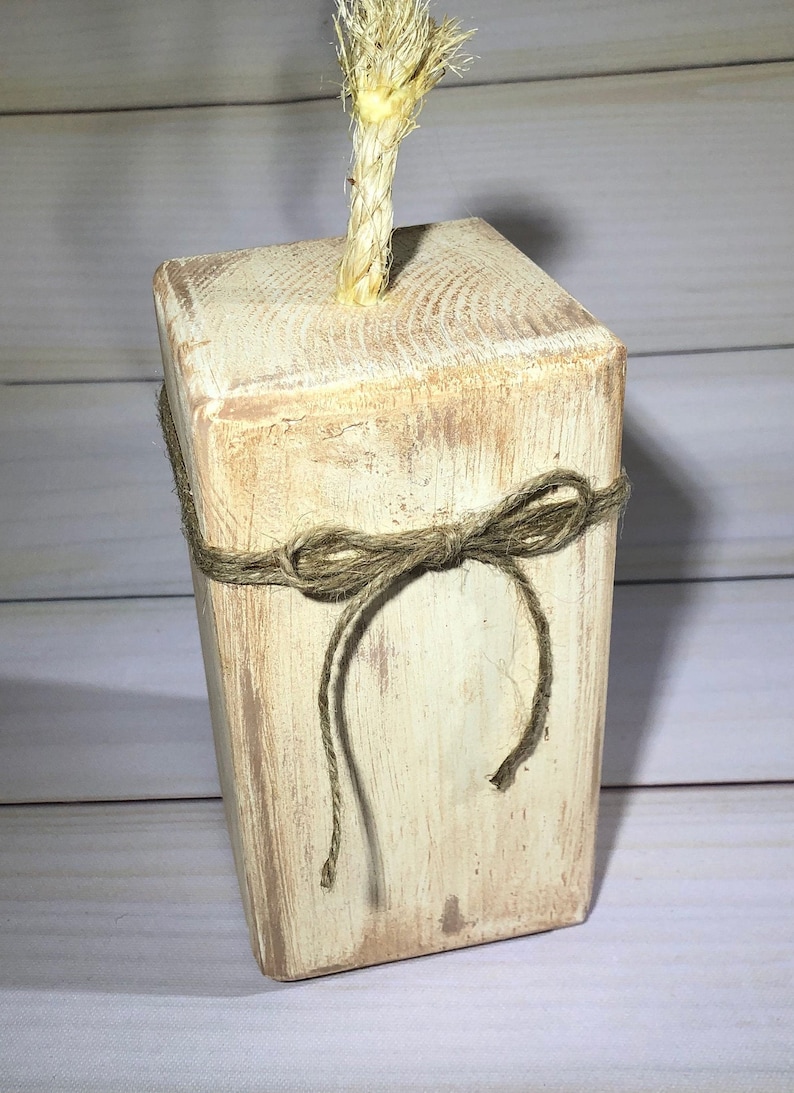 May include: A rustic, rectangular wooden block with a distressed, off-white finish. A natural rope bow is tied around the middle, and a twisted rope extends from the top. The block has visible wood grain.