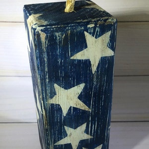 May include: A wooden block painted with a distressed navy blue finish and white stars. A natural rope is attached to the top. The patriotic decor item is a decorative accent for the home.