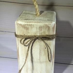 May include: A rustic, rectangular wooden block with a distressed white finish. A natural rope is tied around the middle and a thicker rope protrudes from the top. This decorative piece has a farmhouse aesthetic.