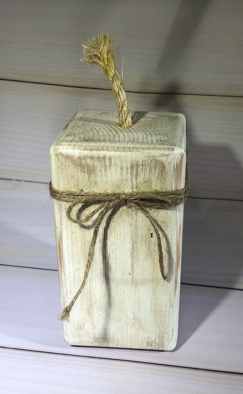 May include: A rustic, rectangular wooden block with a distressed white finish. A natural rope is tied around the middle and a thicker rope protrudes from the top. This decorative piece has a farmhouse aesthetic.